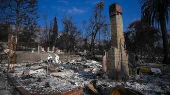 Two people in protective gear inspect the remains of a burned structure surrounded by charred trees and debris.