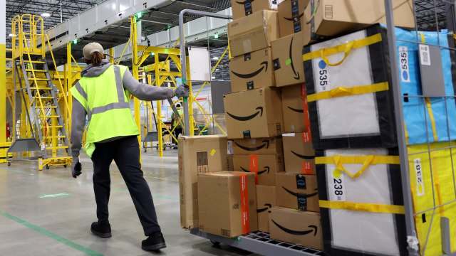 A worker rolls a cart loaded with Amazon packages in a warehouse setting.