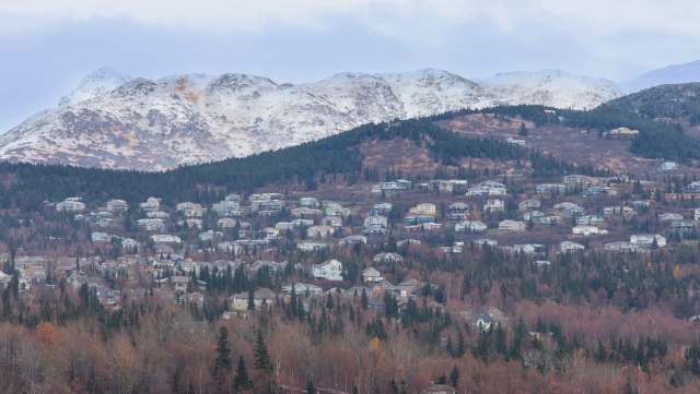 A hillside covered with houses amid trees and mountains with snow-capped peaks in the background.