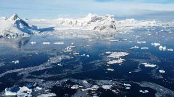 An icy landscape with mountains, floating icebergs, and calm blue water under a clear sky.