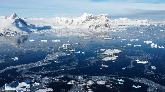 An icy landscape with mountains, floating icebergs, and calm blue water under a clear sky.