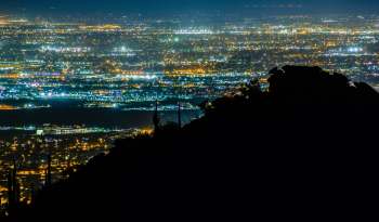 A nighttime cityscape with glittering lights viewed from a mountain silhouette.