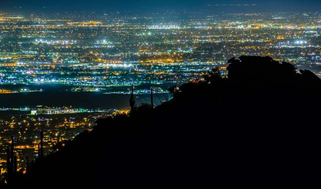 A nighttime cityscape with glittering lights viewed from a mountain silhouette.