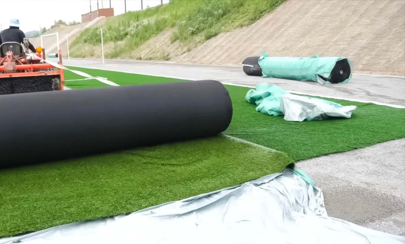 Rolls of artificial turf are being laid out on a sports field with construction materials in the background.