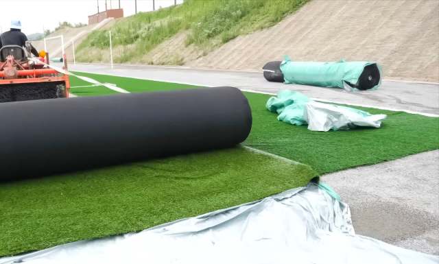 Rolls of artificial turf are being laid out on a sports field with construction materials in the background.