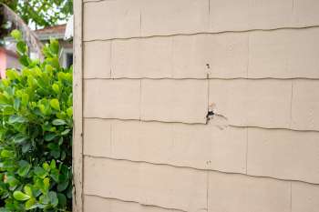 A damaged section of beige siding next to a green shrub.