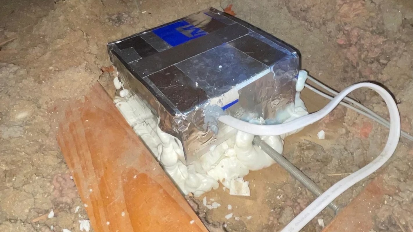 A metal box surrounded by insulation foam and wires on a dirt floor.
