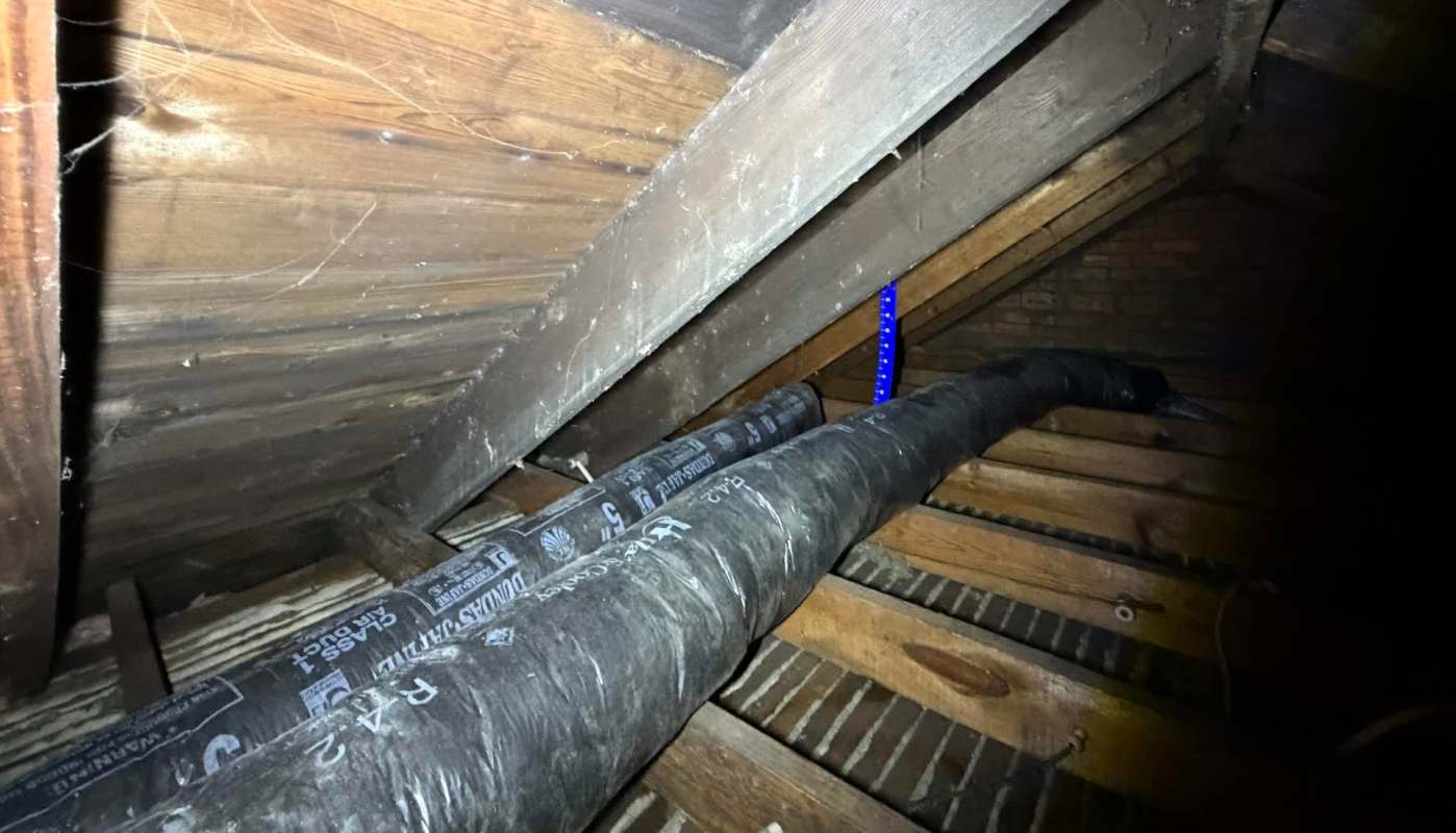 An attic space with wooden beams, insulation, and an insulated duct running along the floor.