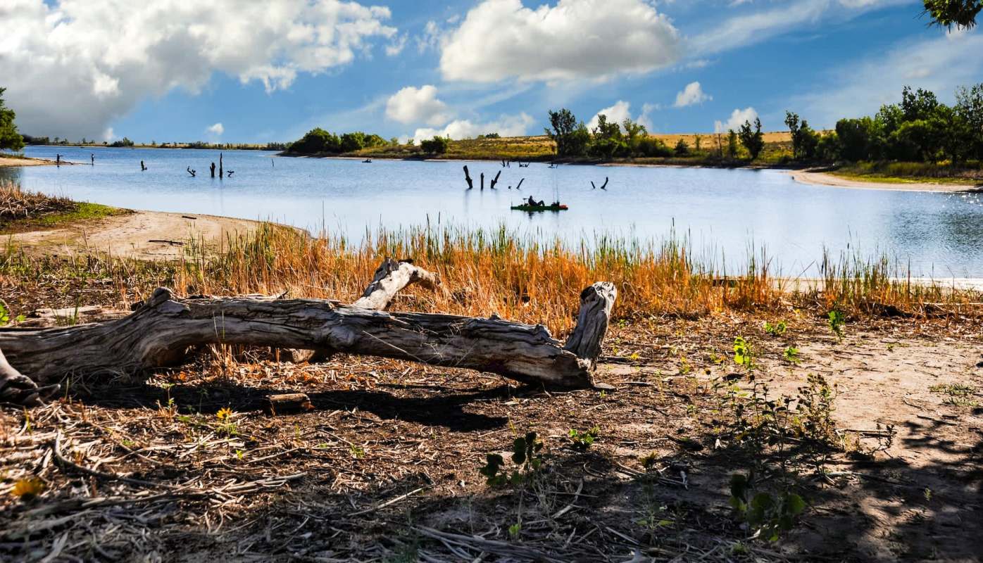 A calm body of water bordered by sandy banks and dry grass, with a lone kayak visible in the distance.
