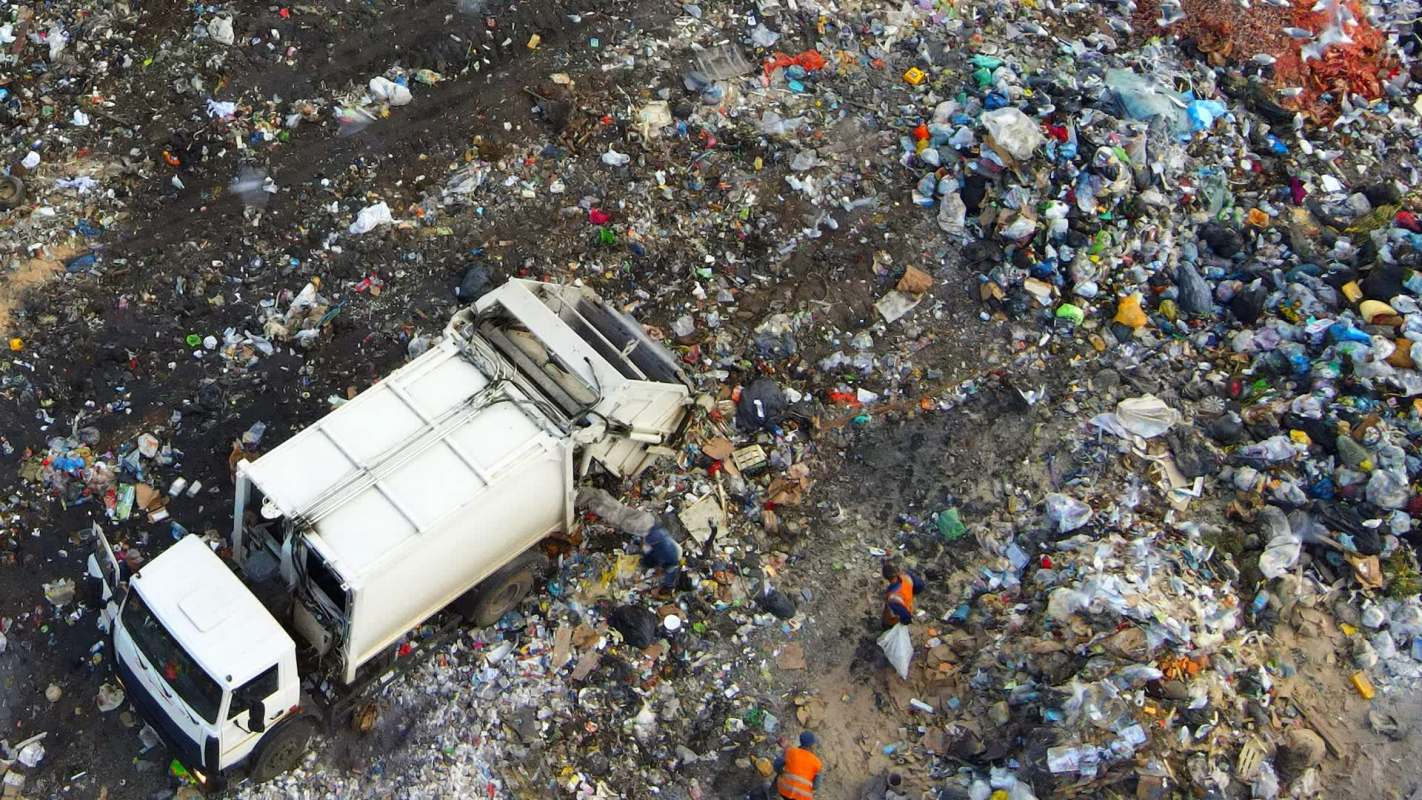 A garbage truck is on a waste pile, with workers collecting debris in a landfill area.