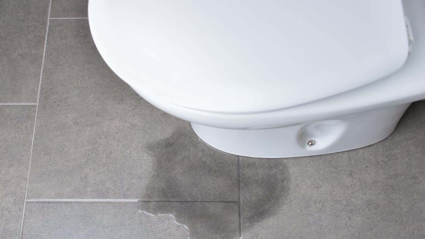 A toilet with water pooling on the gray tiled floor.