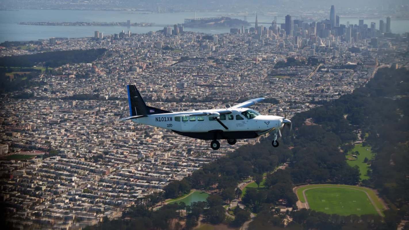 A small airplane flies over a cityscape with buildings and greenery.