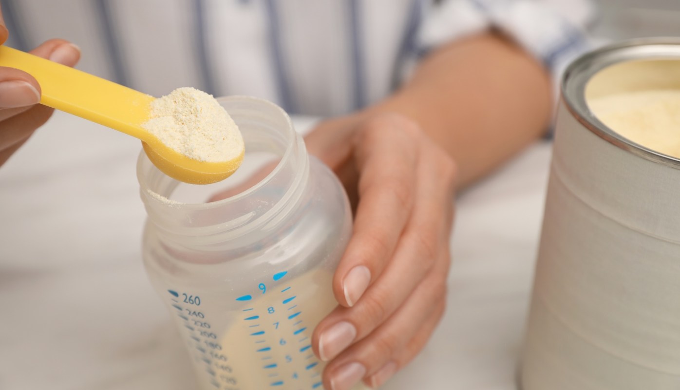 A hand holding a yellow scoop of powder above a baby bottle.