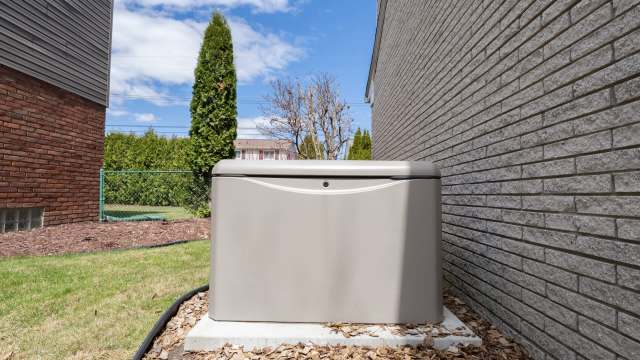 A generator sits on a concrete base next to a brick wall and landscaped yard.