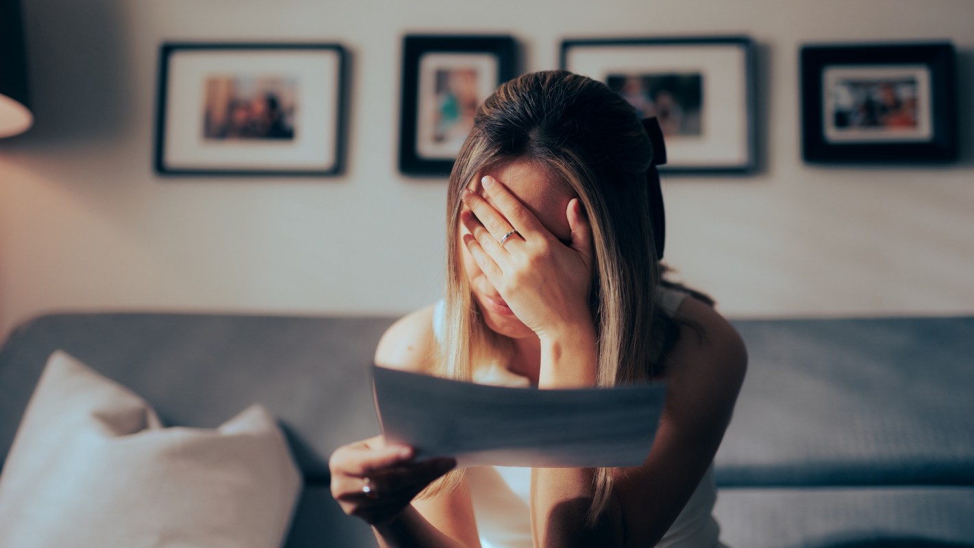 A woman sits on a sofa, holding a piece of paper and covering her face with her hand.