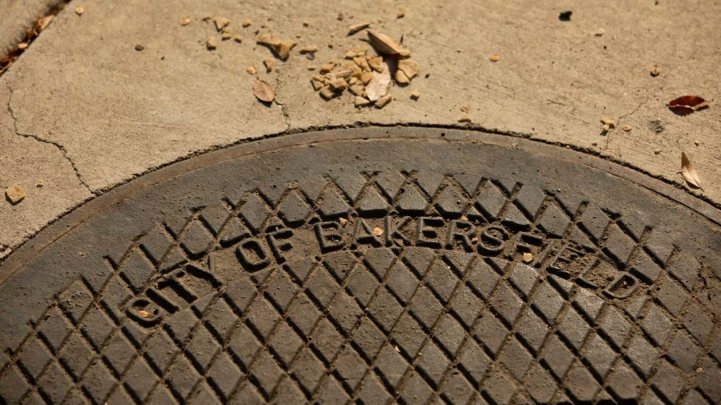 A manhole cover inscribed with "City of Bakersfield" on a concrete surface with scattered leaves and debris.