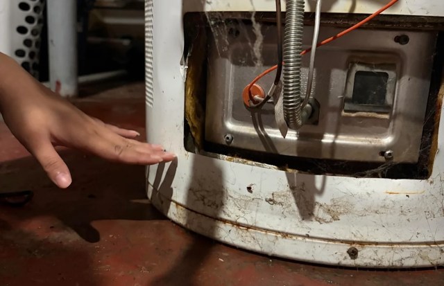 A hand reaches toward a damaged appliance showing wires and dirt near an exposed section.
