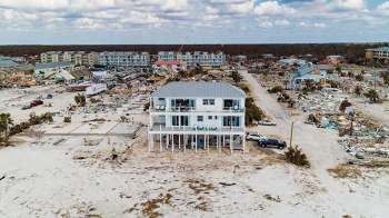 A two-story house stands amid a landscape of debris and damaged buildings.
