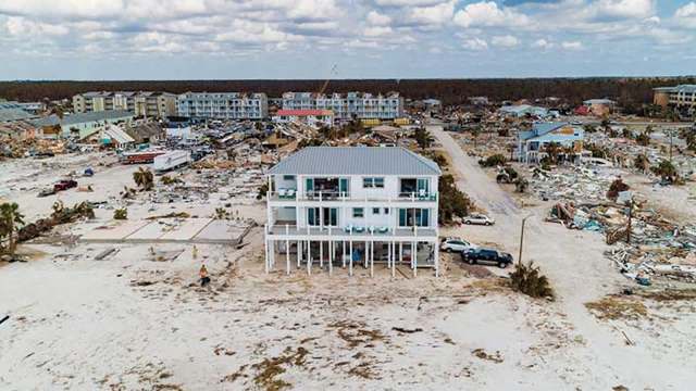 A two-story house stands amid a landscape of debris and damaged buildings.