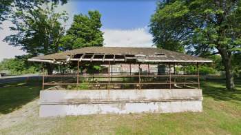An old, partially roofed structure with a concrete base in a grassy area surrounded by trees with an abandoned school in the background.