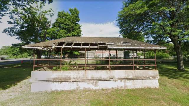 An old, partially roofed structure with a concrete base in a grassy area surrounded by trees with an abandoned school in the background.