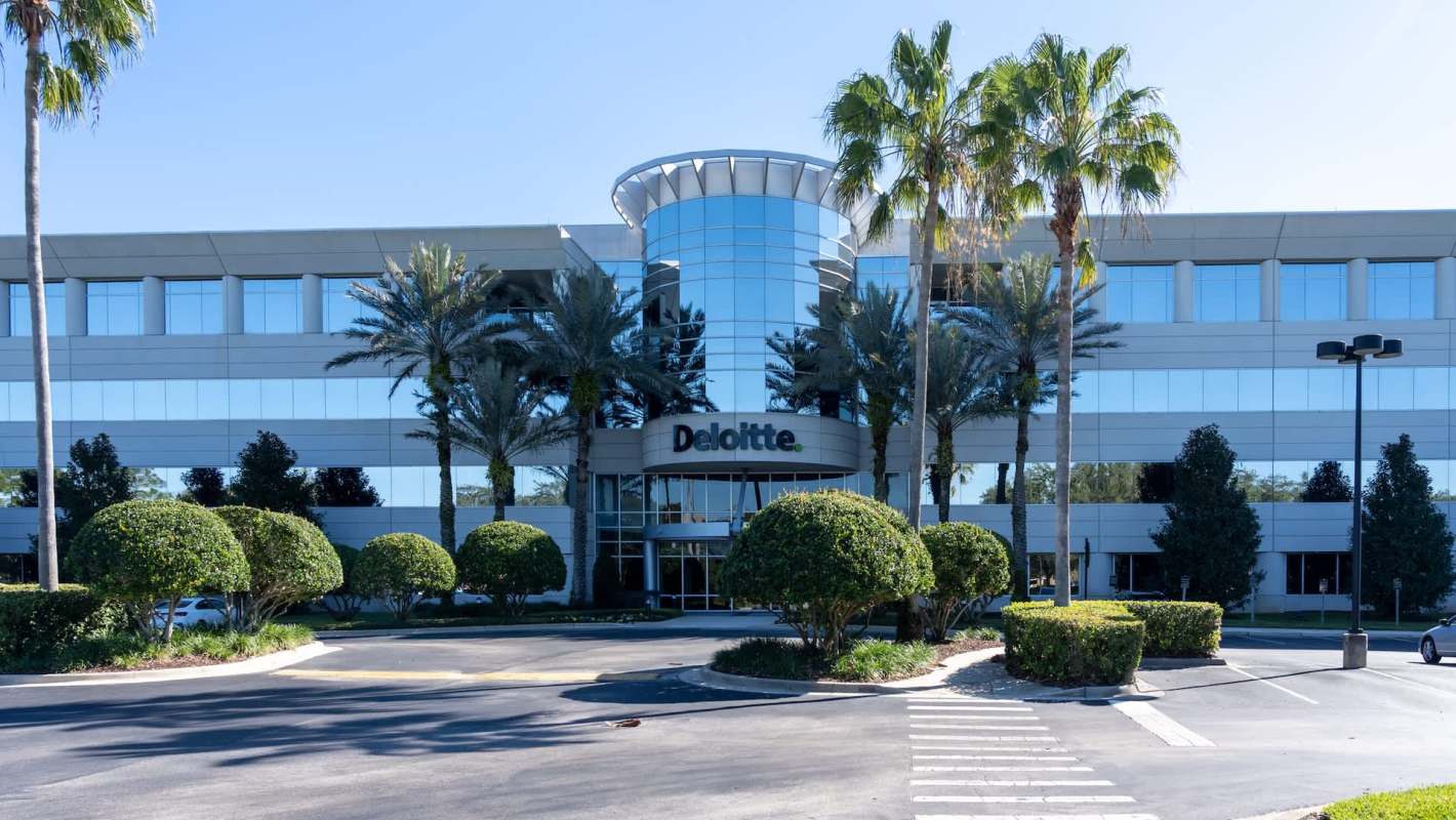 An office building with the Deloitte logo, surrounded by palm trees and manicured hedges.