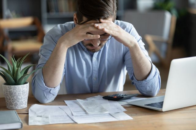 A man sits at a table with bills, a calculator, and a laptop, looking down with his hands on his forehead visibly stressed.