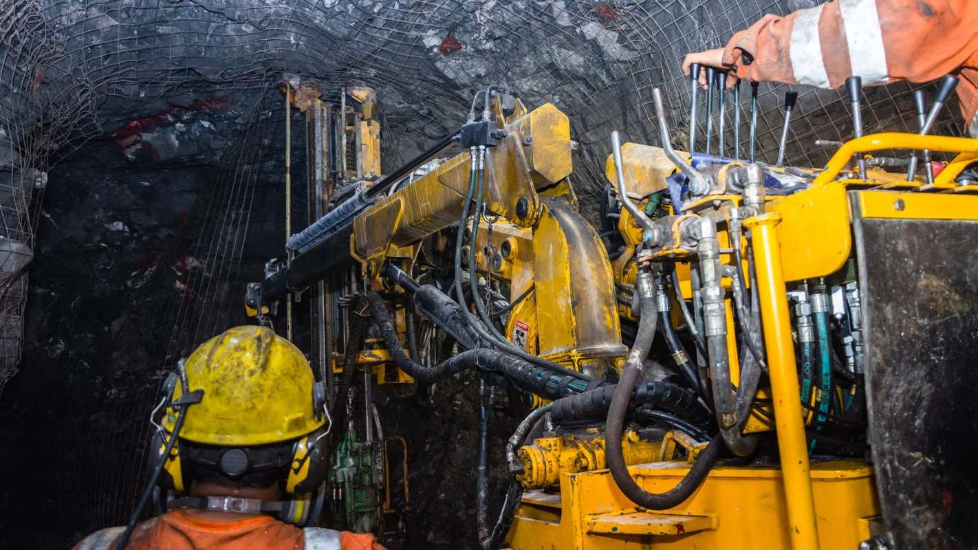 A mining machine operates in an underground tunnel with a worker observing the equipment.