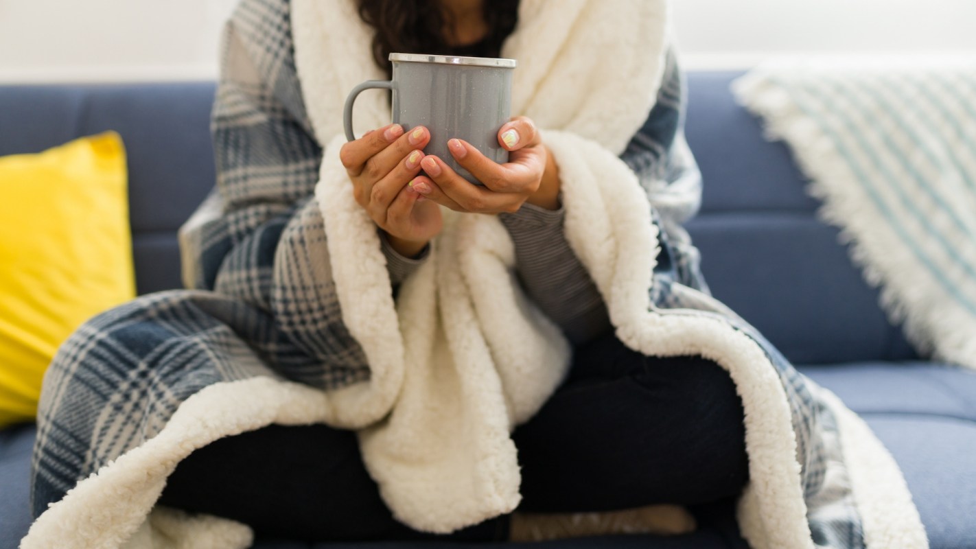 A person wrapped in a blanket holds a mug while sitting on a couch.