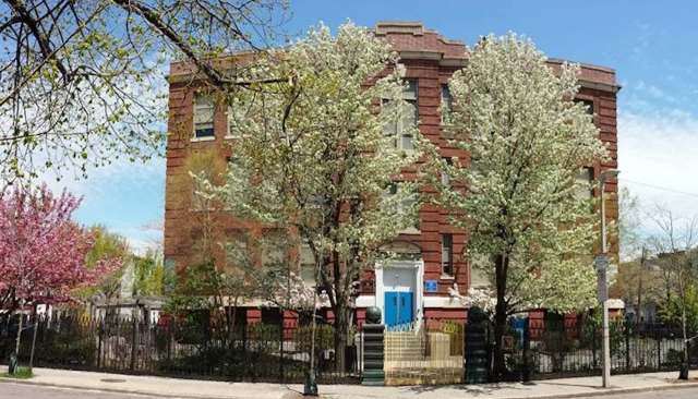 A brick building surrounded by flowering trees and a black iron fence on a clear day.