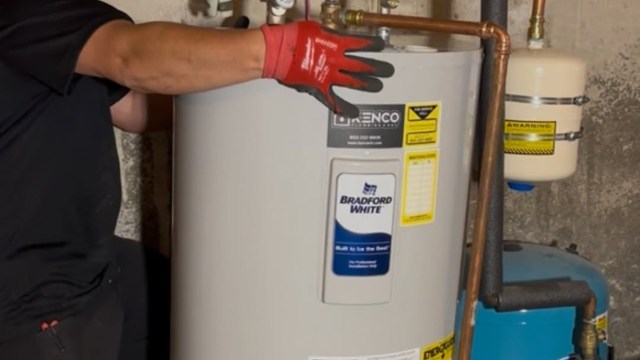 A person in red gloves adjusts a water heater in a basement.