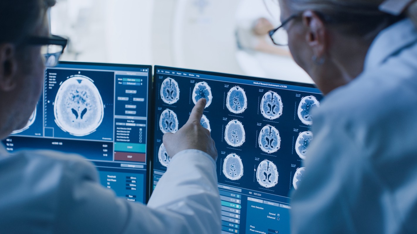 Two medical professionals examine brain scan images on a computer monitor.