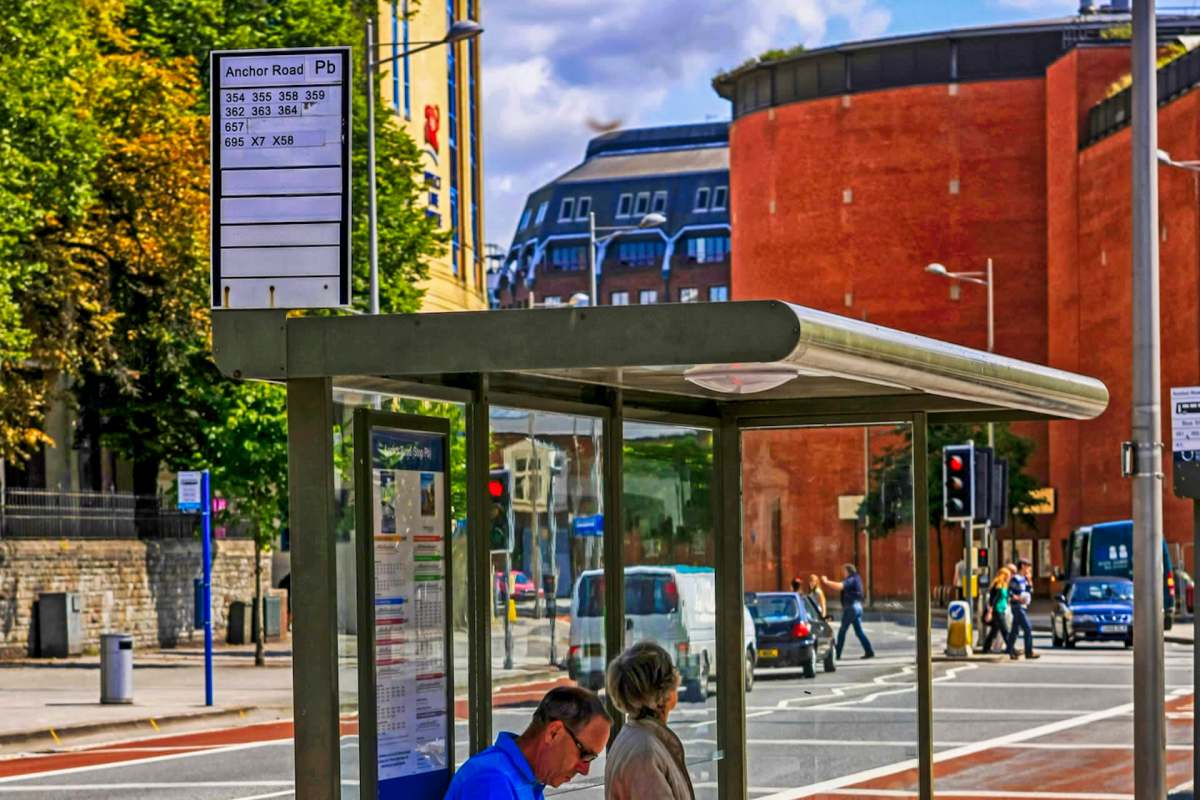 A bus stop with a schedule sign, shelter, and people crossing the street in Bristol, England.
