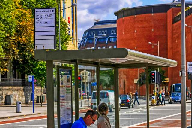 A bus stop with a schedule sign, shelter, and people crossing the street in Bristol, England.