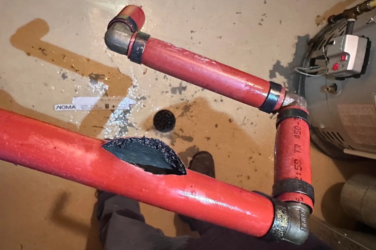 A damaged red pipe with a visible crack, leaking water onto the ground.