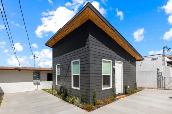 A modern gray tiny house with a peaked roof, surrounded by a driveway and small plants.