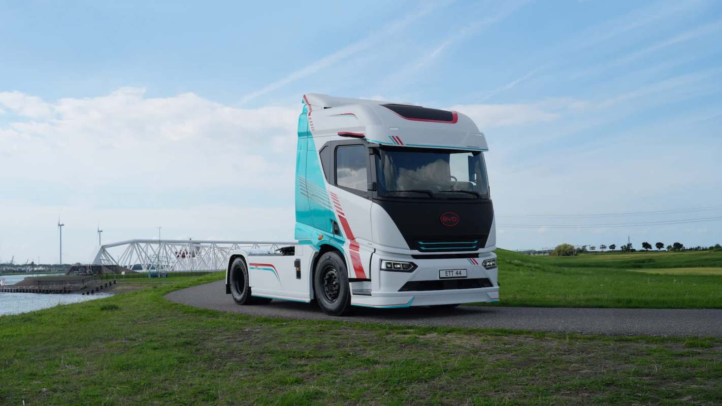 A modern truck parked on a grassy area near a body of water with wind turbines in the background.