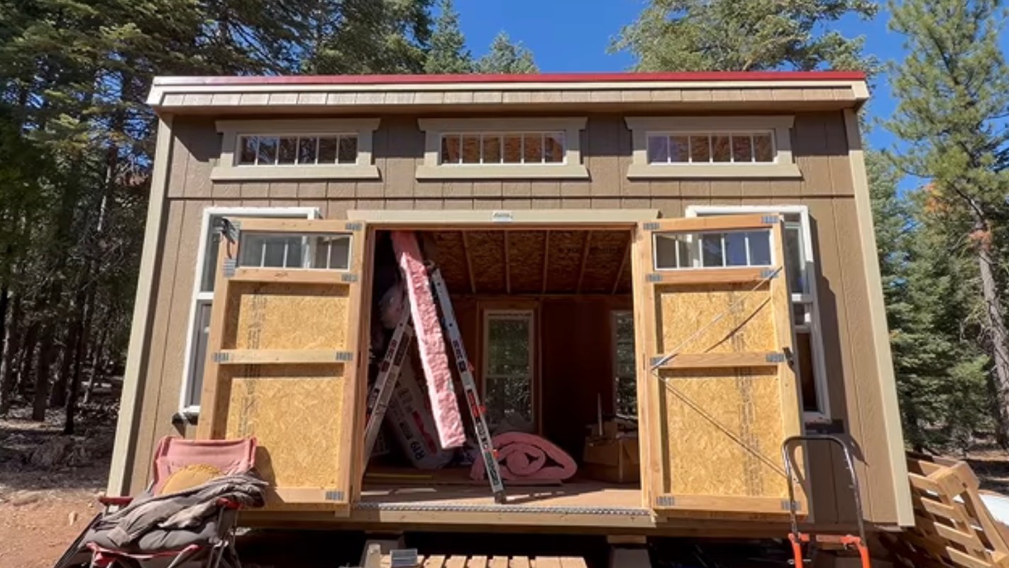 A tiny house with open double doors, exposing interior construction materials and a ladder.