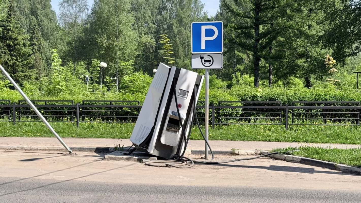 A broken, tilted electric vehicle charging station next to a parking sign in a green area.
