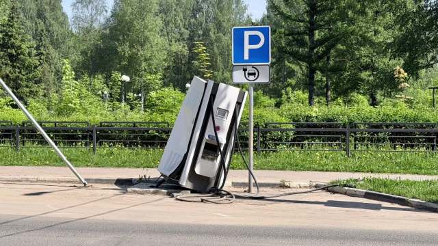 A broken, tilted electric vehicle charging station next to a parking sign in a green area.