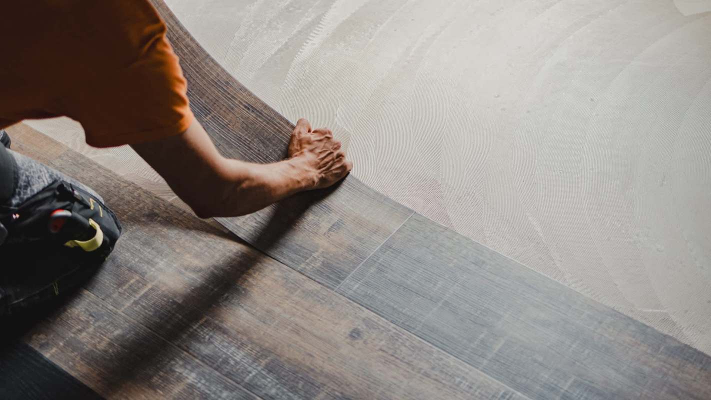 A person is installing flooring, placing a wood plank next to a light-colored surface.