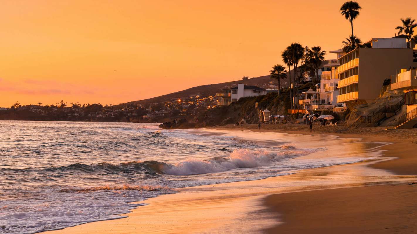 A beach at sunset with gentle waves and palm trees lining the shore alongside coastal buildings.