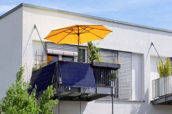 A balcony with solar panels, potted plants, and a large yellow umbrella against a white building.