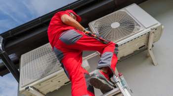 A technician climbs a ladder to service an air conditioning unit mounted on a wall.