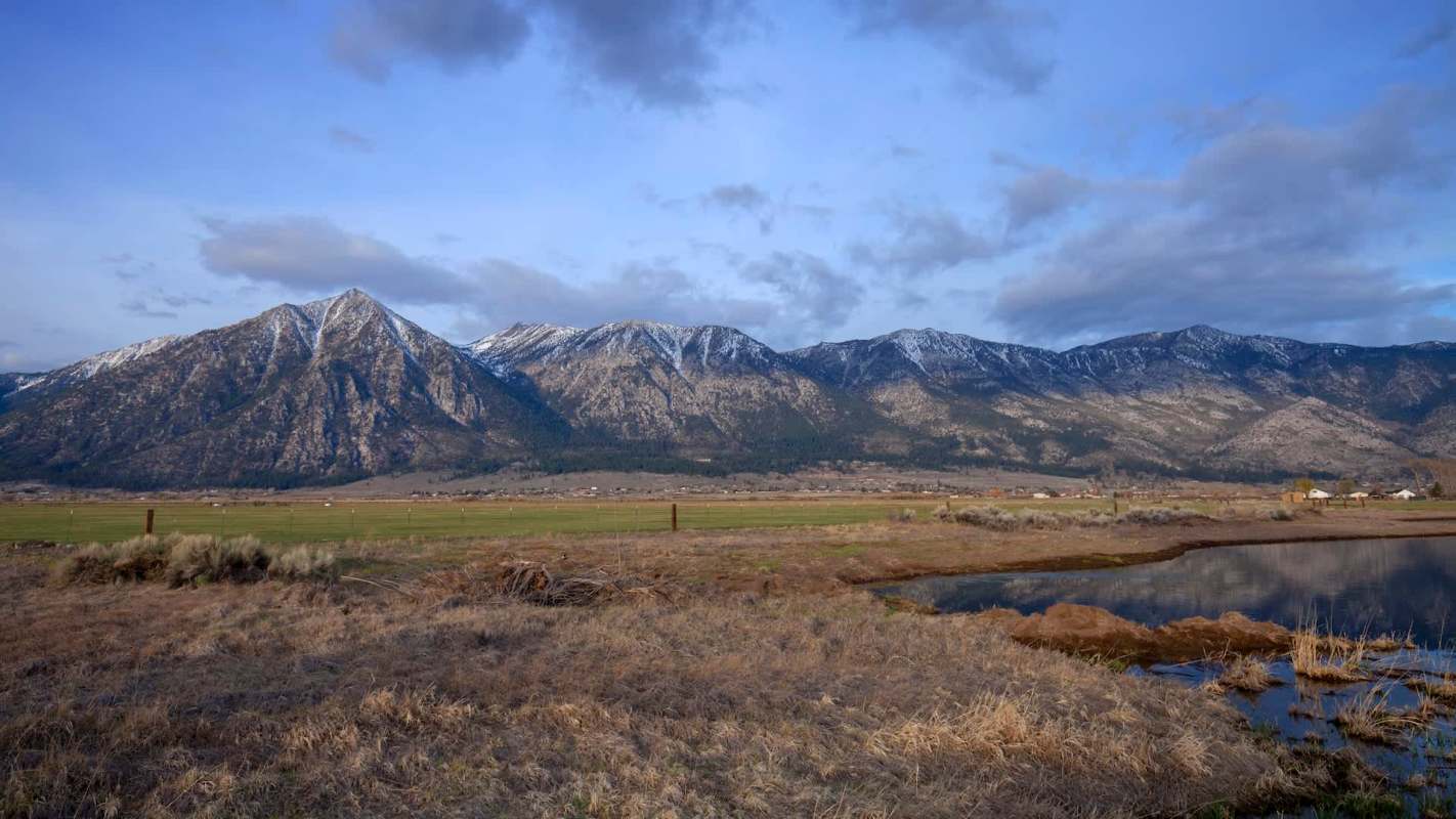 A mountain range under a partly cloudy sky with a grassy field and small pond in the foreground.