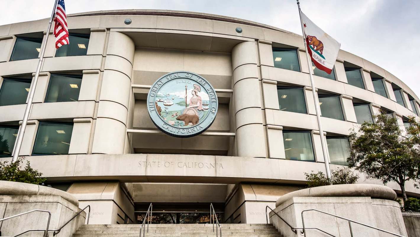 The exterior of a government building featuring the Great Seal of California and American flags.