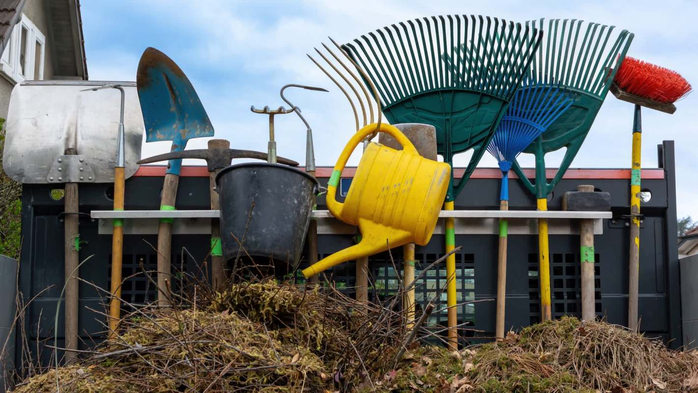 A collection of gardening tools, including shovels, rakes, a watering can, and a bucket outside of a house.