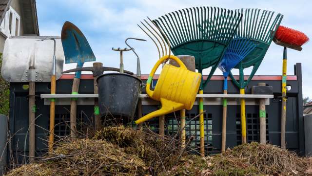 A collection of gardening tools, including shovels, rakes, a watering can, and a bucket outside of a house.