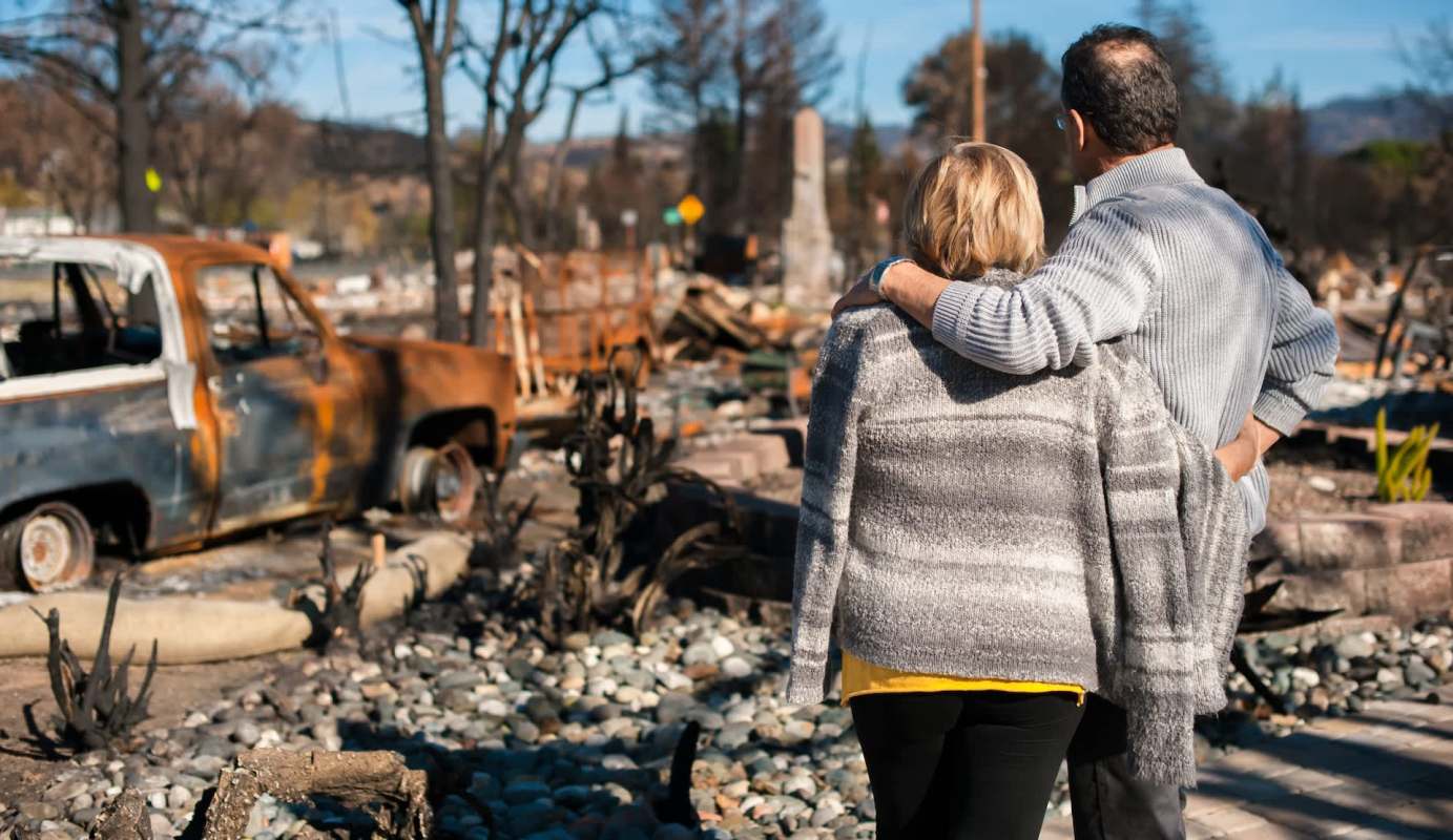 A couple stands together overlooking a burned landscape with a wrecked truck in the foreground.