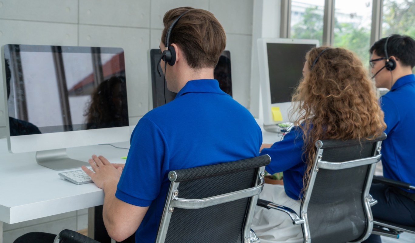 Three individuals wearing headsets are working at computers in a bright office space.